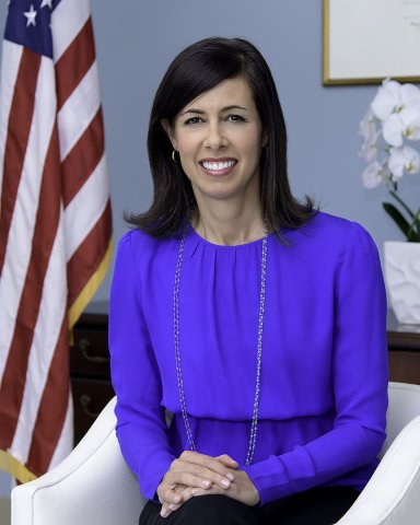 Jessica Rosenworcel sits in a white chair in font of an American flag. She has long, straight, brown hair and wears a thin gold chain. 