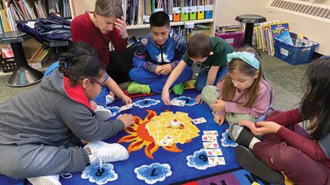 A group of students and their teacher play a board game on a rug in the classroom