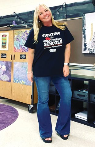 Blond teacher stands in a classroom wearing a black t-shirts that says Fighting for Great Public Schools