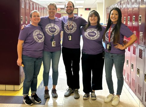 5 teachers wearing matching purple pro-union t-shirts stand in a school hallway lined with lockers