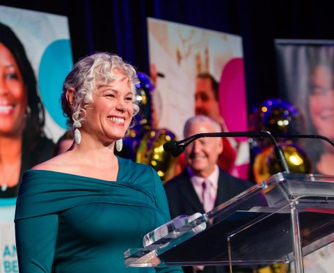 Photo of Jen Bramson, a woman with silver hair and a teal dress smiling and standing at a podium