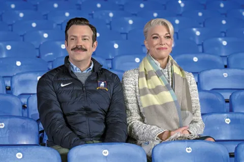 Jason Sudeikis as Ted Lasso with Hannah Waddingham as Rebecca Welton sitting in blue stadium seats on the show Ted Lasso.