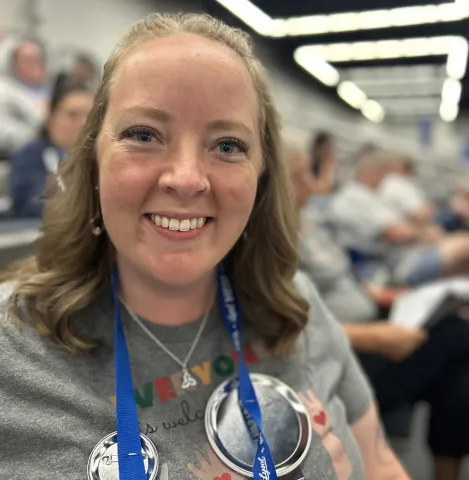 A woman proudly displaying a blue badge on her t-shirt, with a friendly smile directed at the camera.