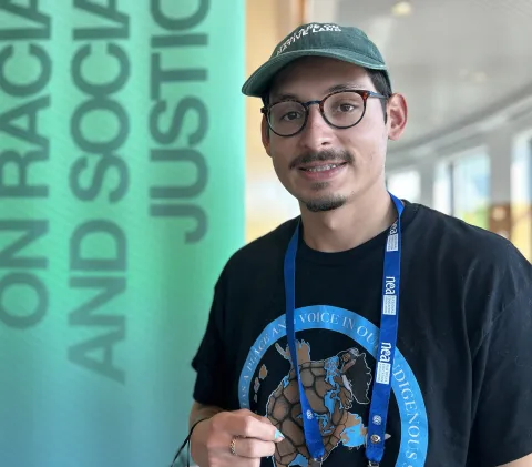 A man in a hat and glasses stands smiling in front of a green banner.