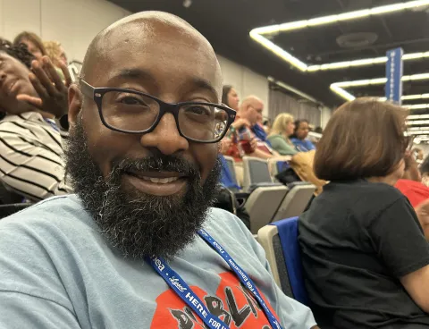 A thoughtful man with a beard and glasses sits in a room, surrounded by other people.